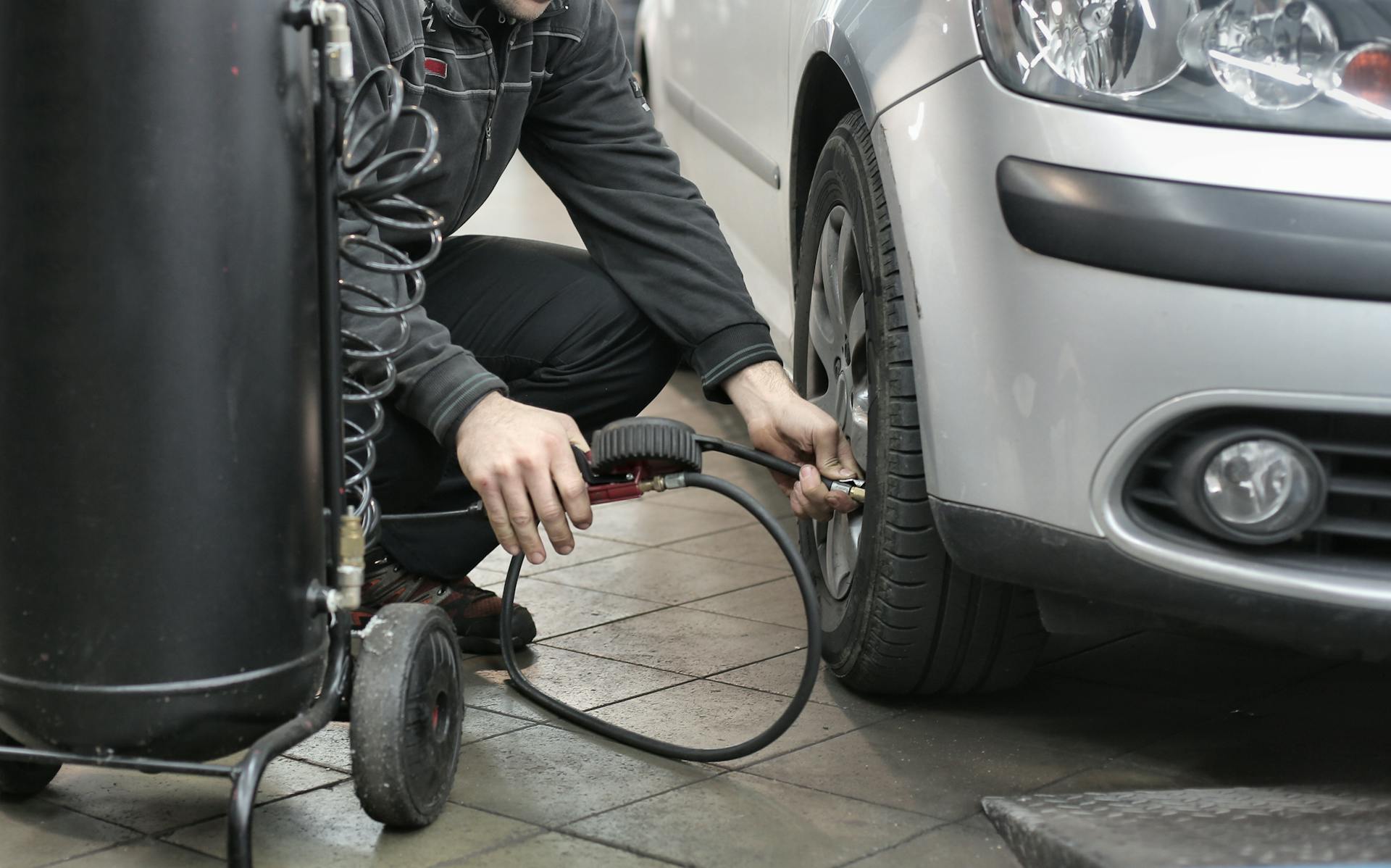 Mechanic inflating the front right tire of a silver car using an air compressor hose in a garage.